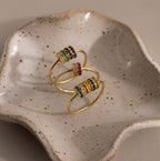 Two Multiple Birthstone Bead Rings, stackable and set with colorful gems, rest on a speckled ceramic dish with wavy edges.