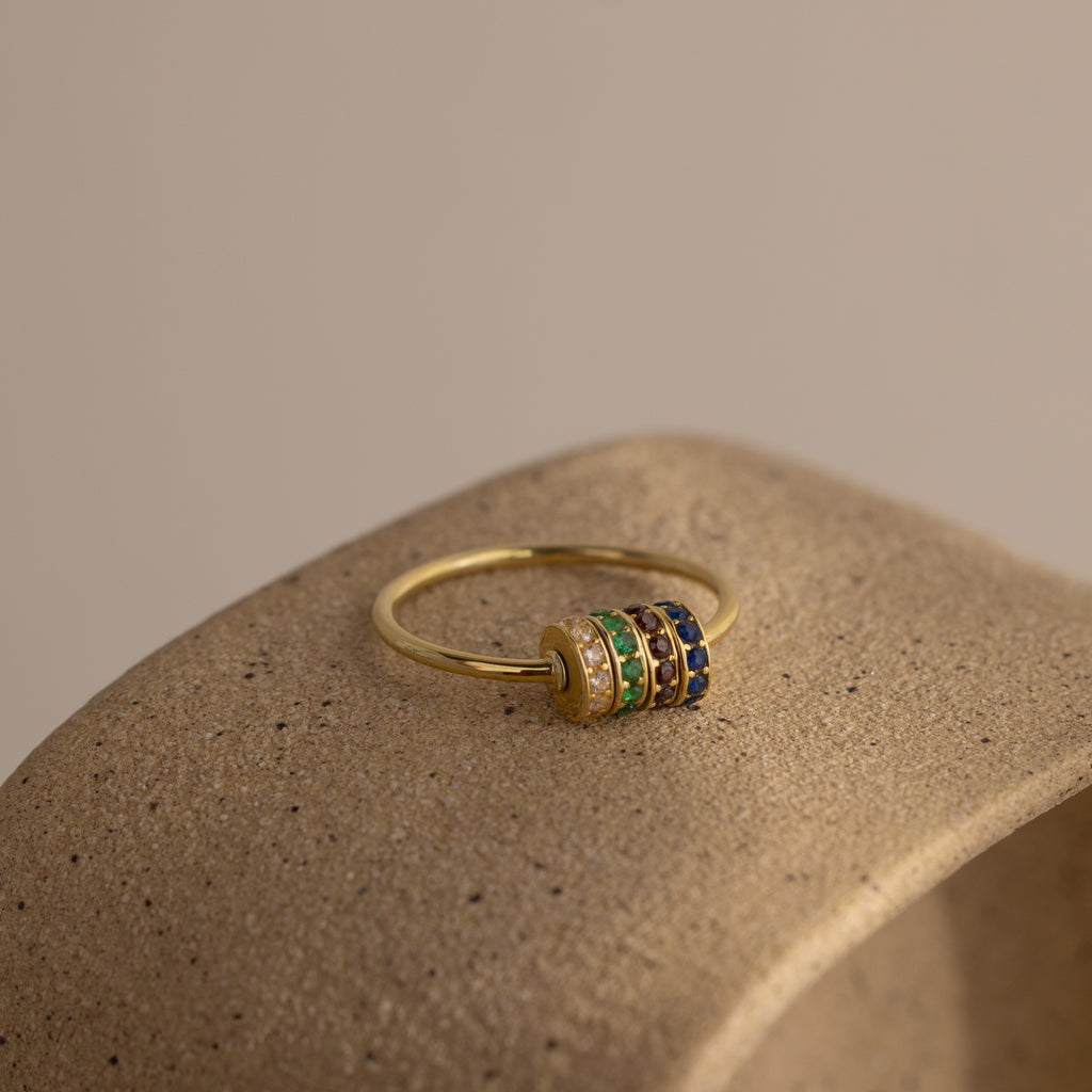 The Multiple Birthstone Bead Ring in 18K Gold features multicolored gemstone bands, perfect as a personalized birthstone ring or stackable jewelry, shown on a textured beige stone surface.