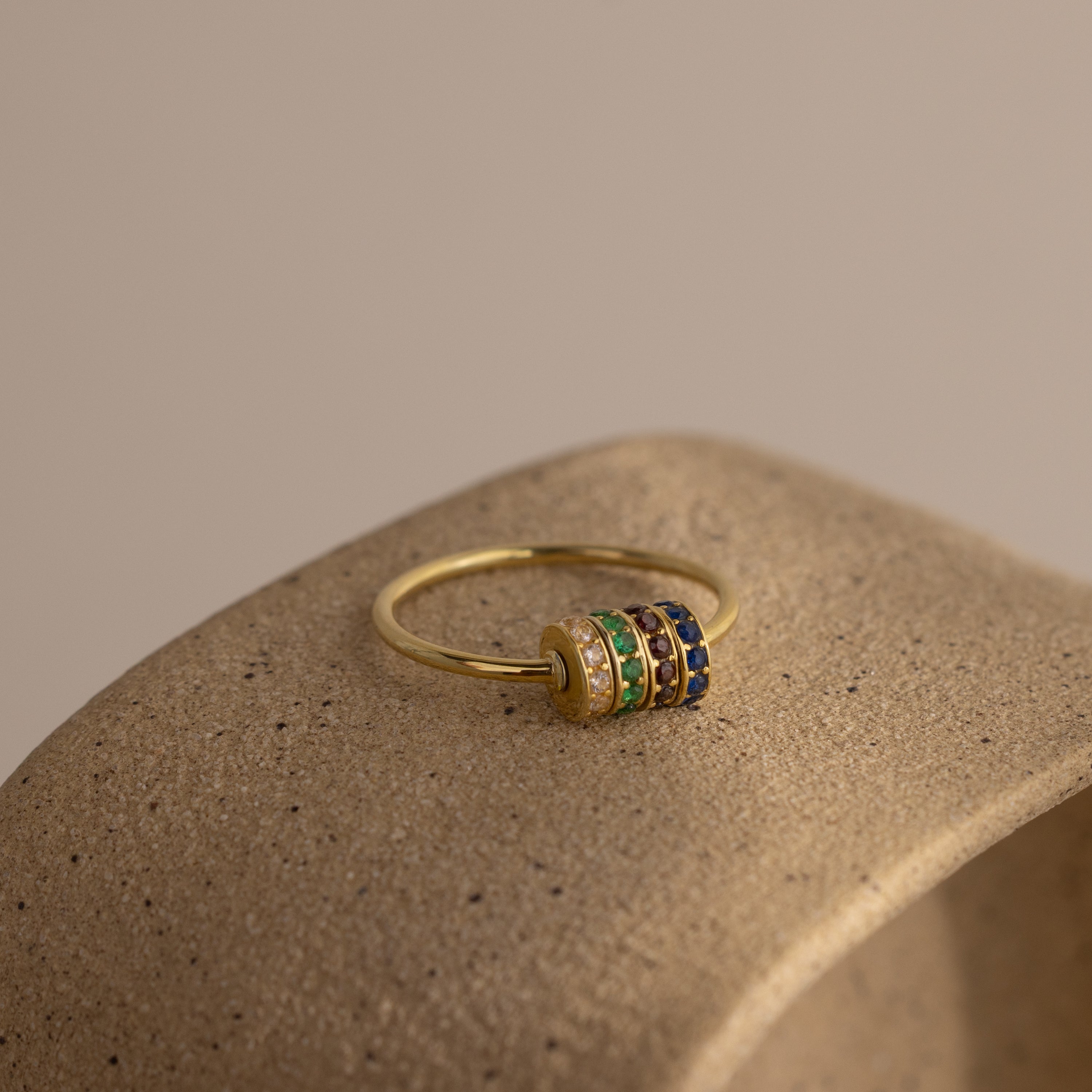 The Multiple Birthstone Bead Ring in 18K Gold features multicolored gemstone bands, perfect as a personalized birthstone ring or stackable jewelry, shown on a textured beige stone surface.