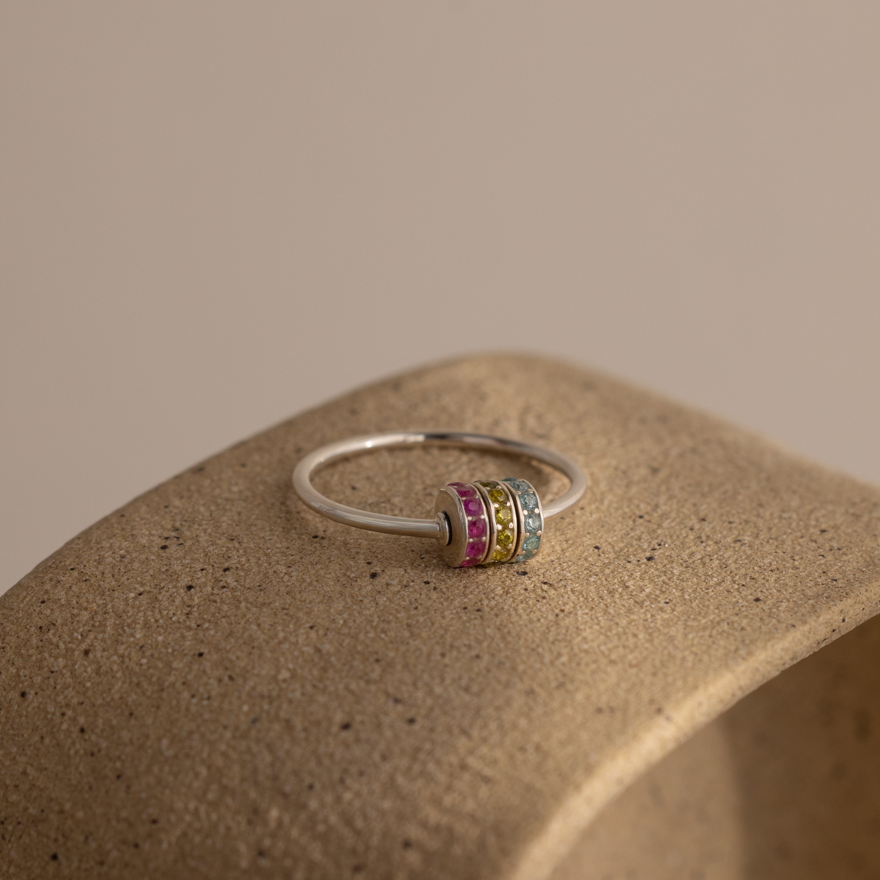 The Multiple Birthstone Bead Ring in Sterling Silver, featuring vibrant gemstone bands, sits atop a textured beige surface—a perfect choice for personalized jewelry.