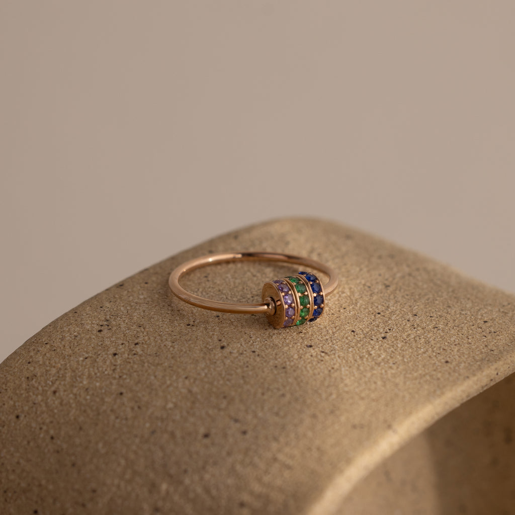 The Multiple Birthstone Bead Ring in Rose Gold features five colorful gemstone bands and sits on a textured beige surface, making it ideal for those who love personalized jewelry.