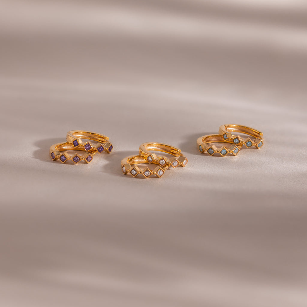 Five pairs of gold rings, including the Geometric Diamond Huggies adorned with diamond accents and small colorful gemstones, are displayed on a soft beige surface.