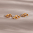 Five pairs of gold rings, including the Geometric Diamond Huggies adorned with diamond accents and small colorful gemstones, are displayed on a soft beige surface.