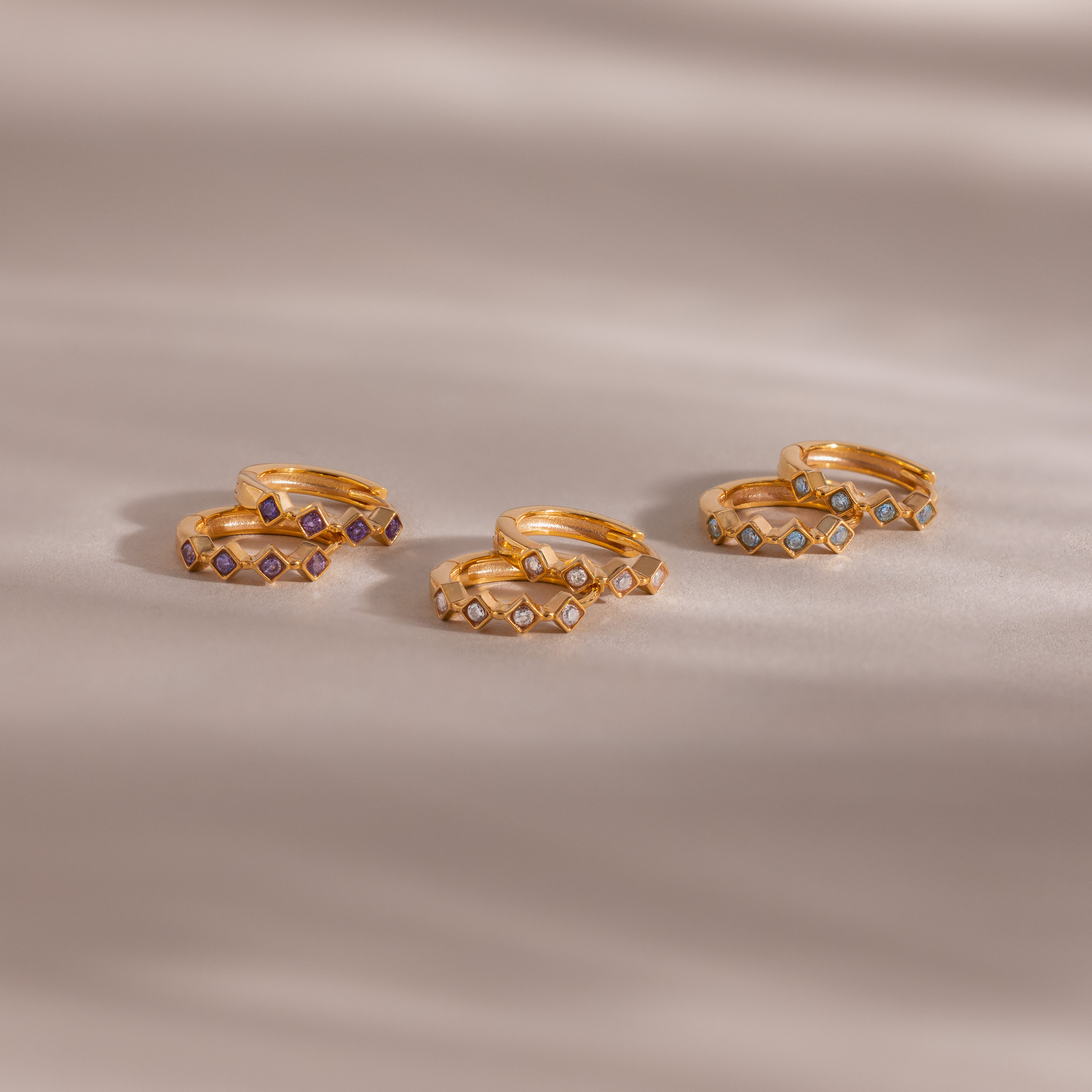 Five pairs of gold rings, including the Geometric Diamond Huggies adorned with diamond accents and small colorful gemstones, are displayed on a soft beige surface.