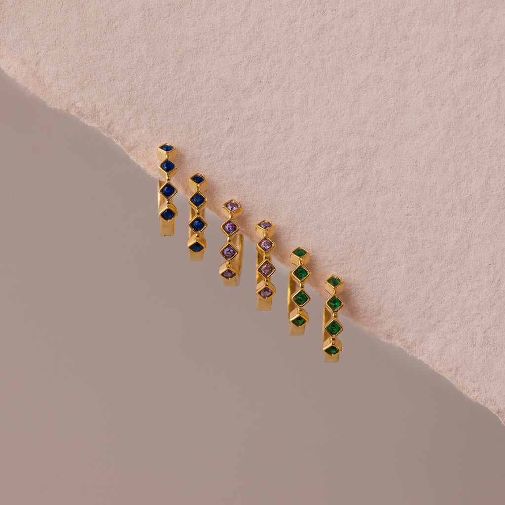 Six pairs of Geometric Sapphire Huggies with blue, purple, and green stones displayed on a textured pink and beige background—ideal for anyone seeking unique, modern birthstone jewelry.