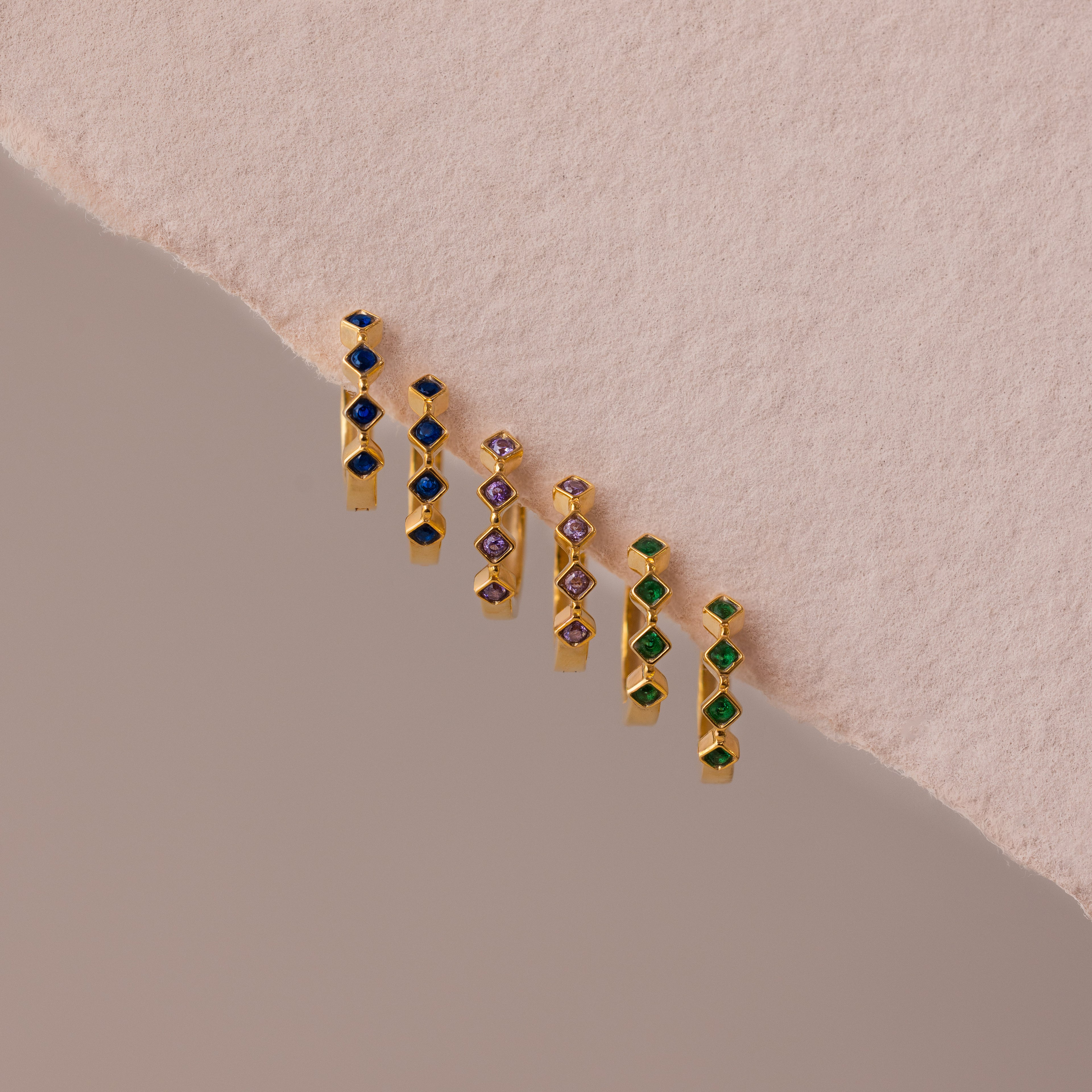 Six pairs of Geometric Sapphire Huggies with blue, purple, and green stones displayed on a textured pink and beige background—ideal for anyone seeking unique, modern birthstone jewelry.