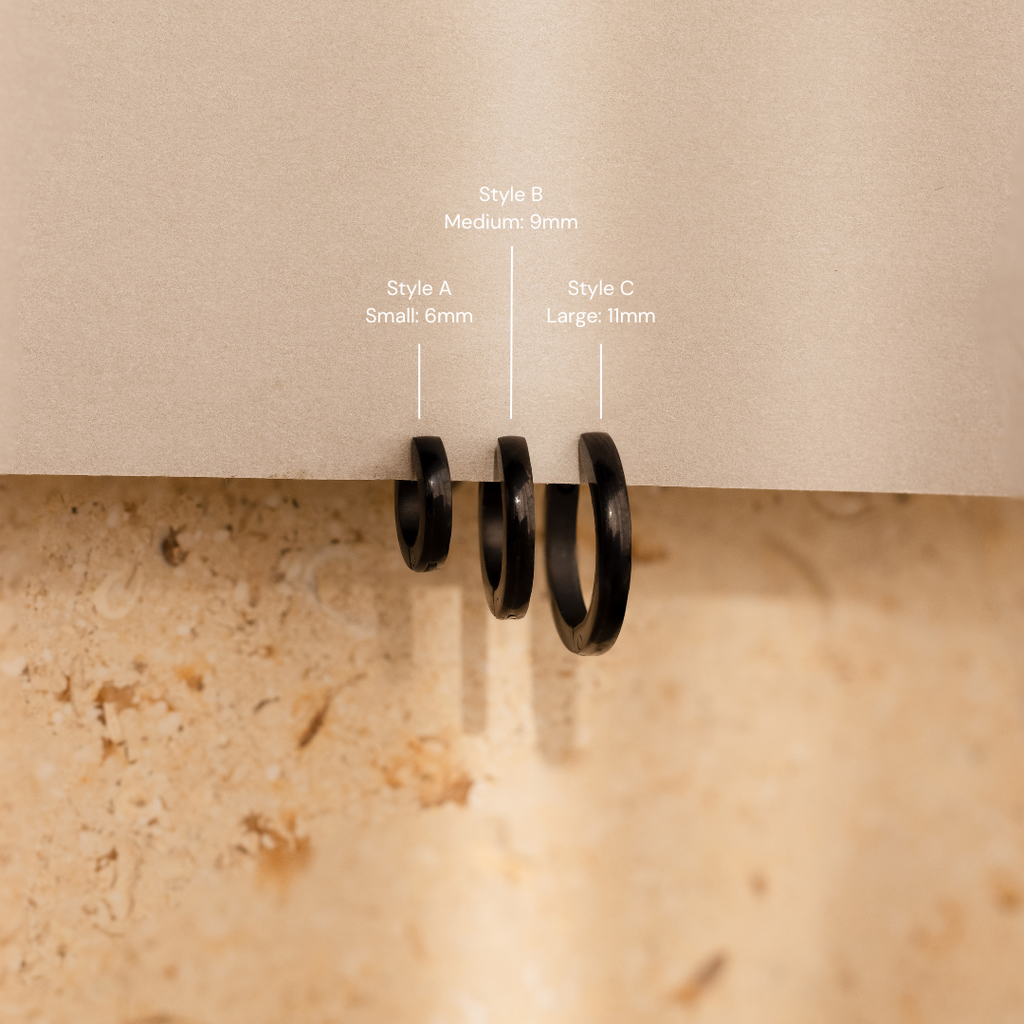 The Men's Black Hoops Set features three sleek black earrings in small, medium, and large sizes, displayed on a beige surface with labels—an ideal stylish gift for him.