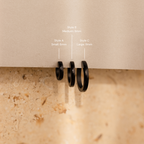 The Men's Black Hoops Set features three sleek black earrings in small, medium, and large sizes, displayed on a beige surface with labels—an ideal stylish gift for him.