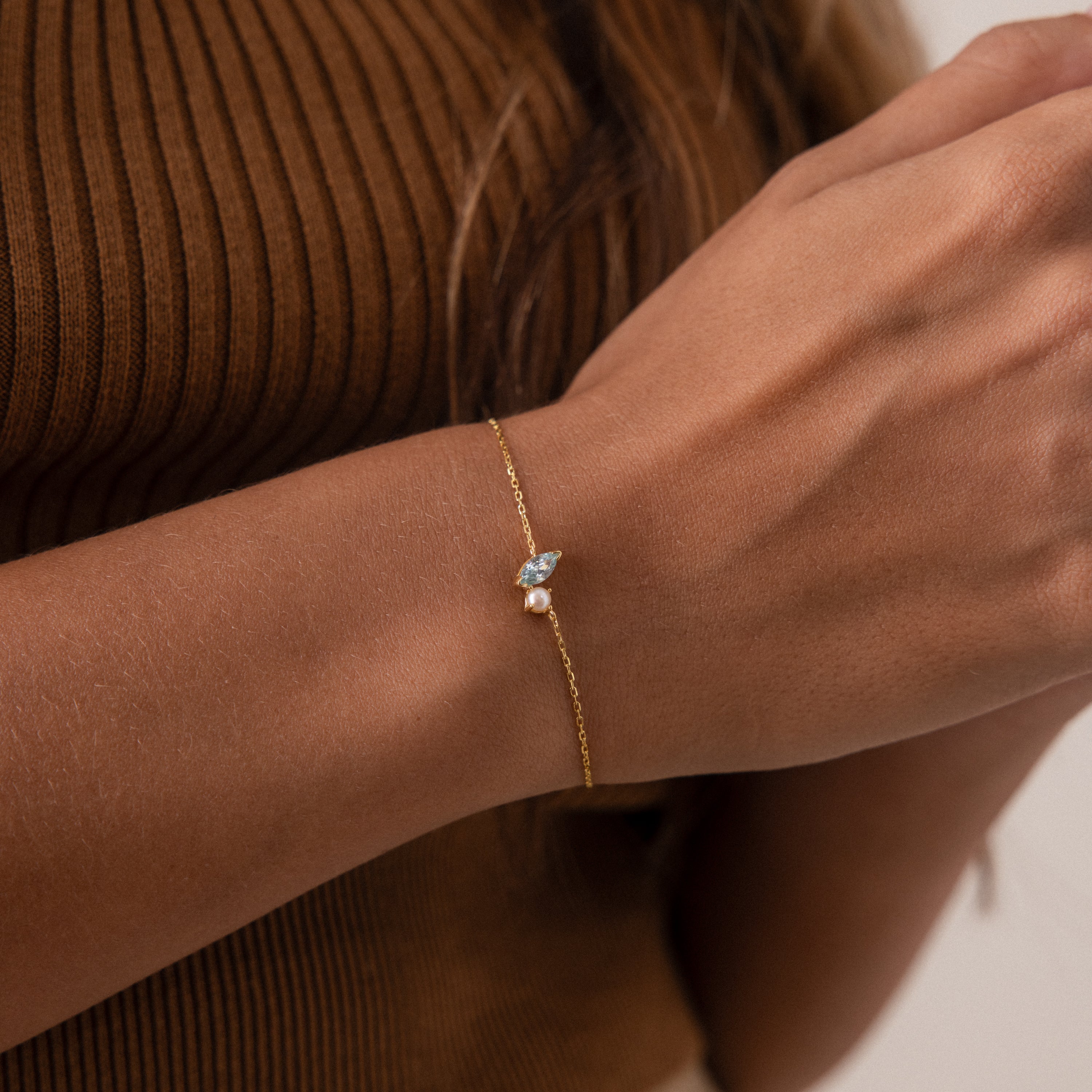 A woman wearing a gold Marquise Blue Zircon & Pearl Bracelet with a small gemstone, dressed in a brown ribbed top.