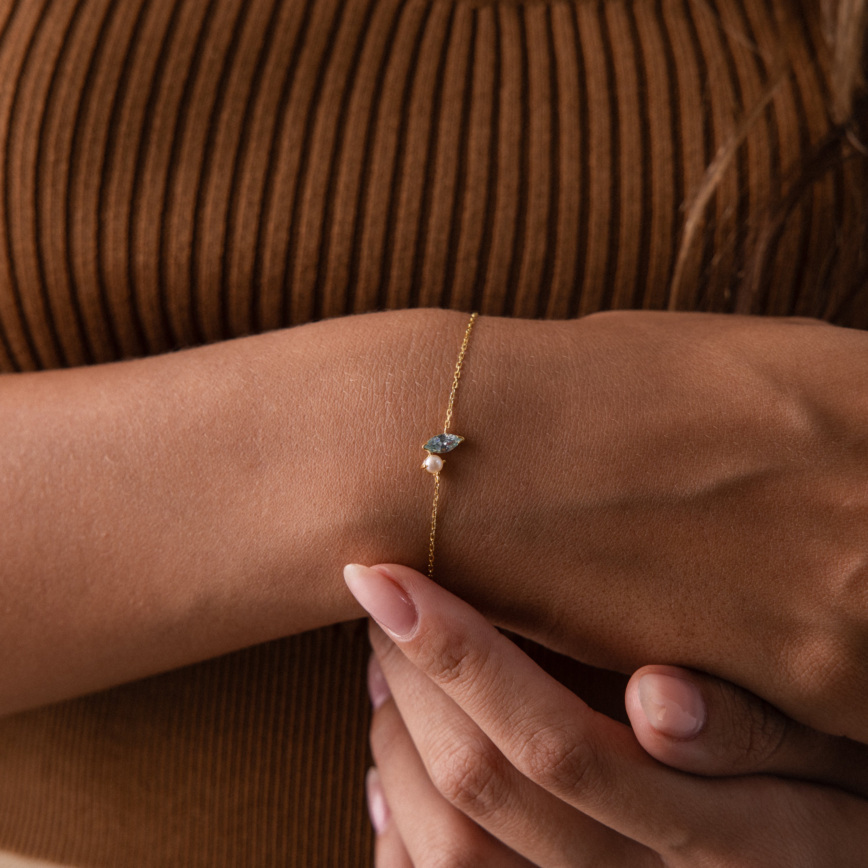 A woman's wrist adorned with the Marquise Birthstone & Pearl Bracelet, featuring a delicate blue stone and pearl, set against a brown ribbed top—a refined and elegant gift for someone special.