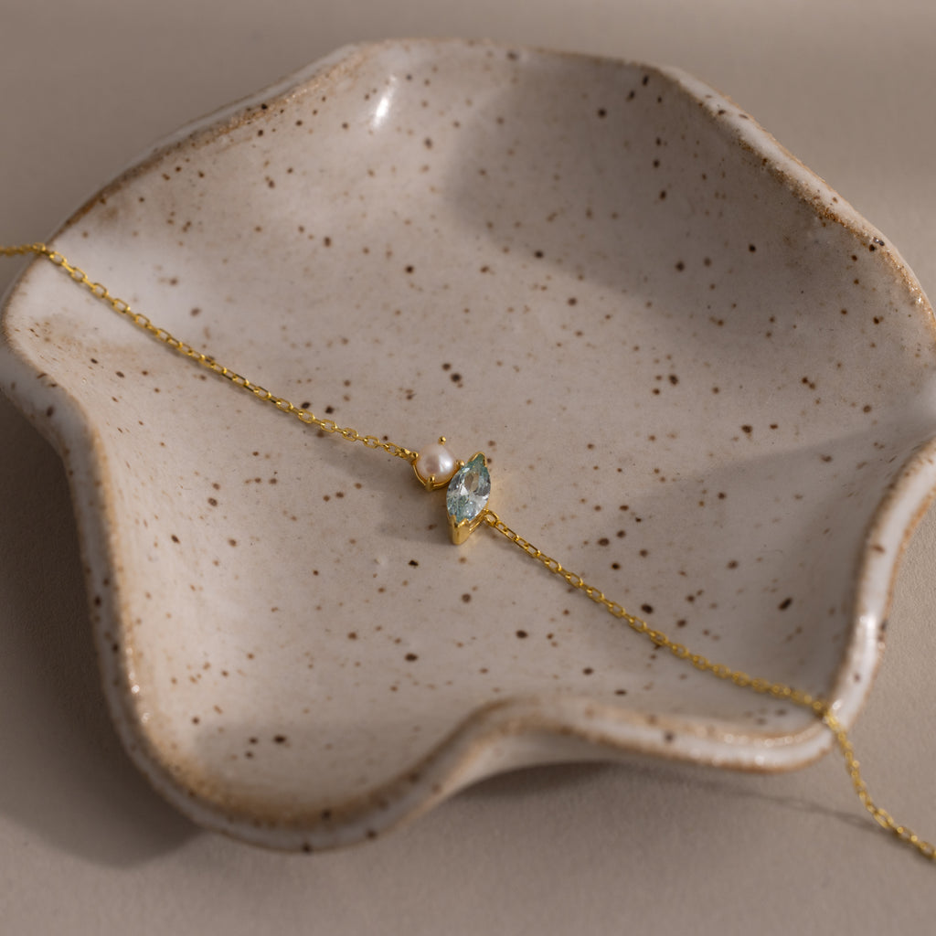 The Marquise Aquamarine & Pearl Bracelet, an elegant gift for someone special, is beautifully displayed on a speckled, wavy ceramic dish.