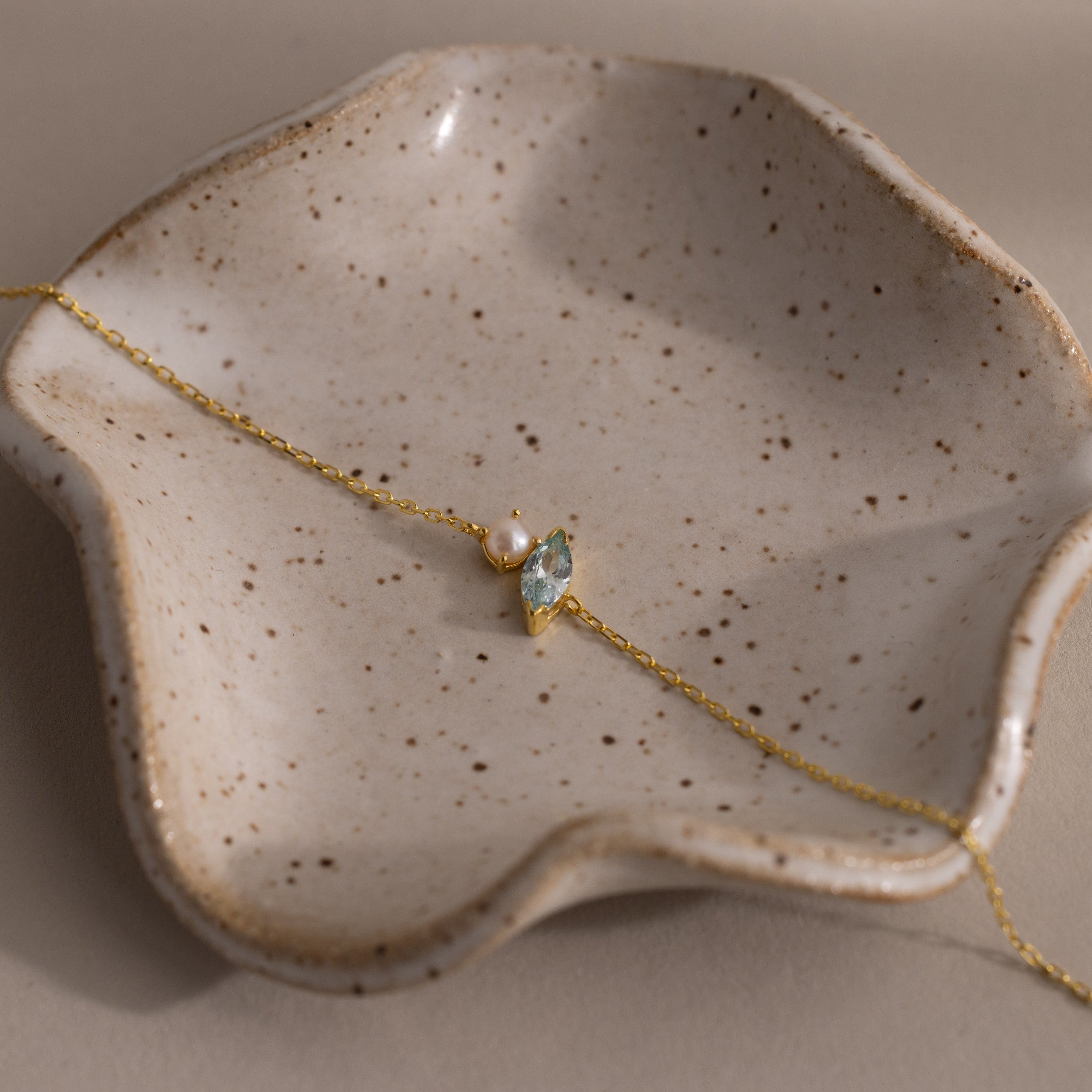 The Marquise Aquamarine & Pearl Bracelet, an elegant gift for someone special, is beautifully displayed on a speckled, wavy ceramic dish.