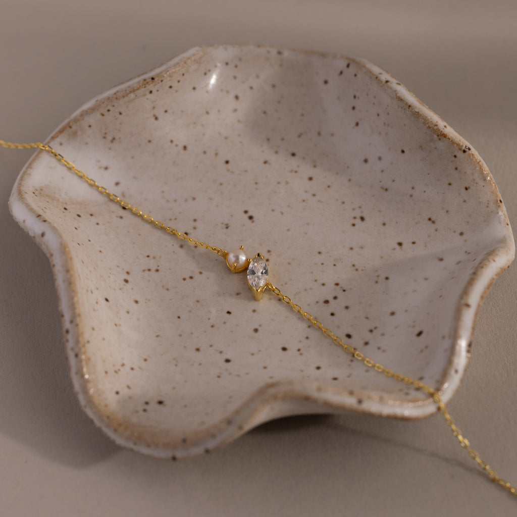 Marquise Diamond & Pearl Bracelet—featuring a pearl and crystal charm on a gold chain—makes a perfect gift, beautifully displayed on a textured, speckled ceramic dish.