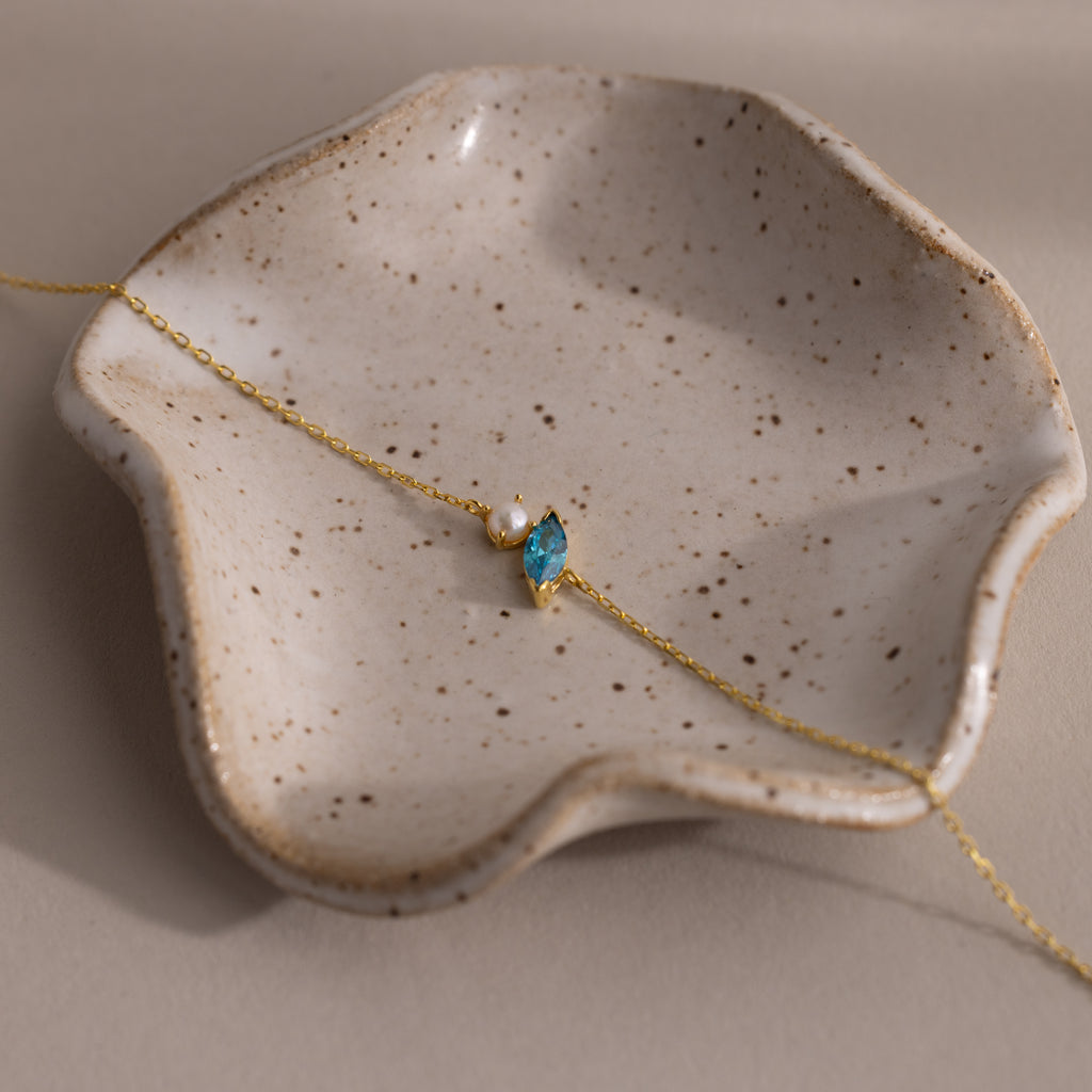 The Marquise Blue Zircon & Pearl Bracelet, featuring a blue gem and pearl, is displayed on a speckled wavy ceramic dish—a perfect gift for someone special.