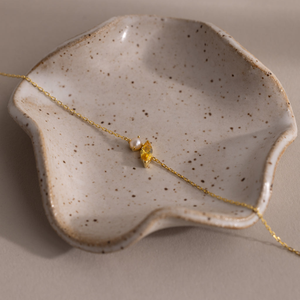 The Marquise Topaz & Pearl Bracelet features a gold chain, pearl accents, and a gold leaf charm—an elegant gift inspired by the timeless beauty of marquise birthstones. Displayed on a speckled ceramic dish.