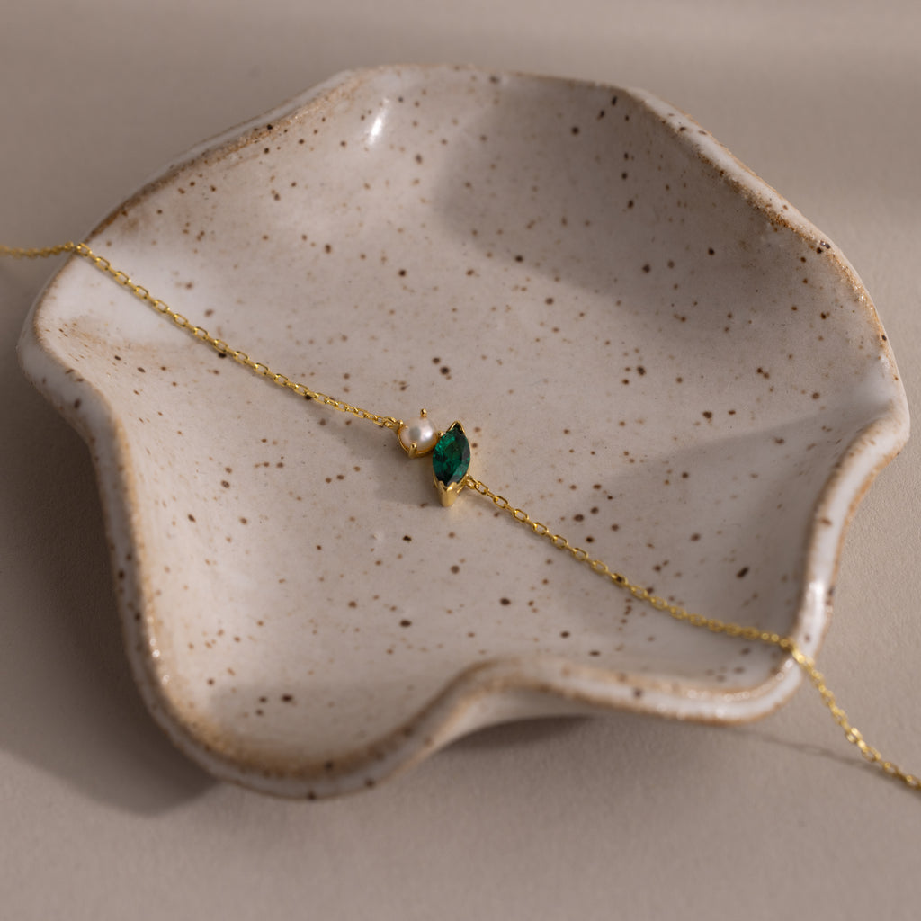 The Marquise Emerald & Pearl Bracelet, featuring a gold chain with green gemstone and pearl, is elegantly displayed on a speckled ceramic dish—making it a perfect gift for someone special.