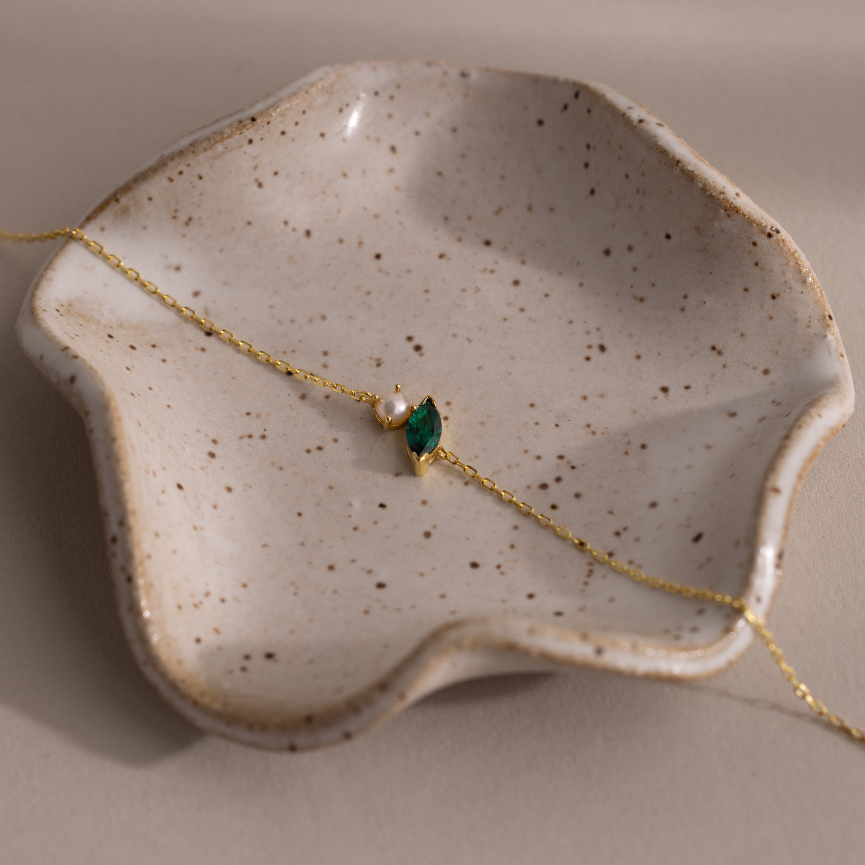 The Marquise Emerald & Pearl Bracelet, featuring a gold chain with green gemstone and pearl, is elegantly displayed on a speckled ceramic dish—making it a perfect gift for someone special.