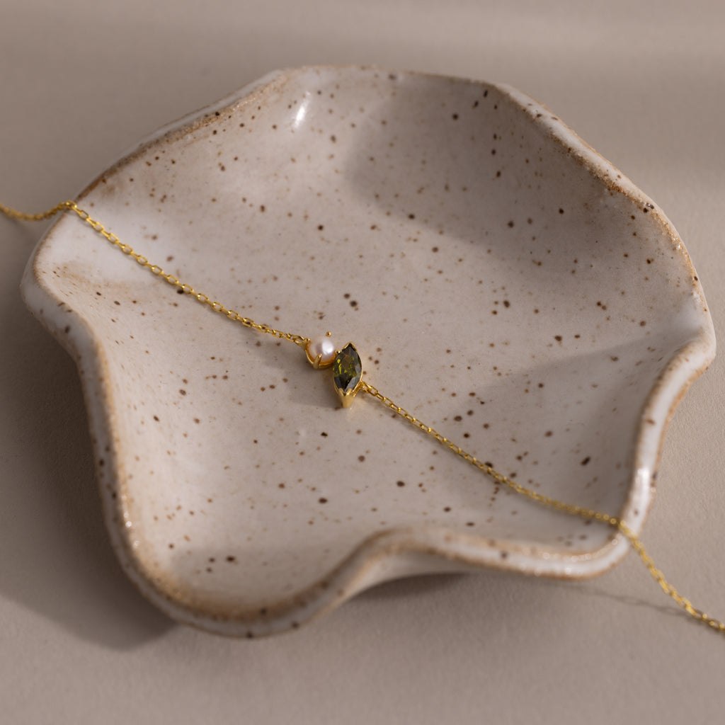 The Marquise Peridot & Pearl Bracelet, featuring a lustrous pearl and green peridot, is elegantly displayed on a wavy-edge ceramic dish—a perfect gift for someone special.