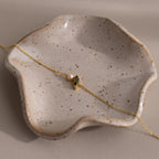 The Marquise Peridot & Pearl Bracelet, featuring a lustrous pearl and green peridot, is elegantly displayed on a wavy-edge ceramic dish—a perfect gift for someone special.