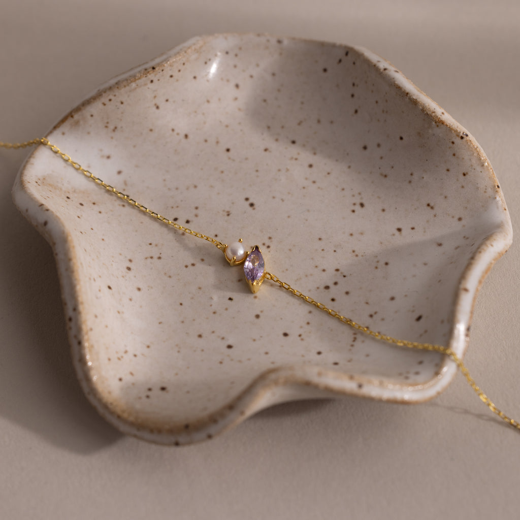 The Marquise Alexandrite & Pearl Bracelet rests on a speckled, wavy ceramic dish—an elegant gift for any special occasion.