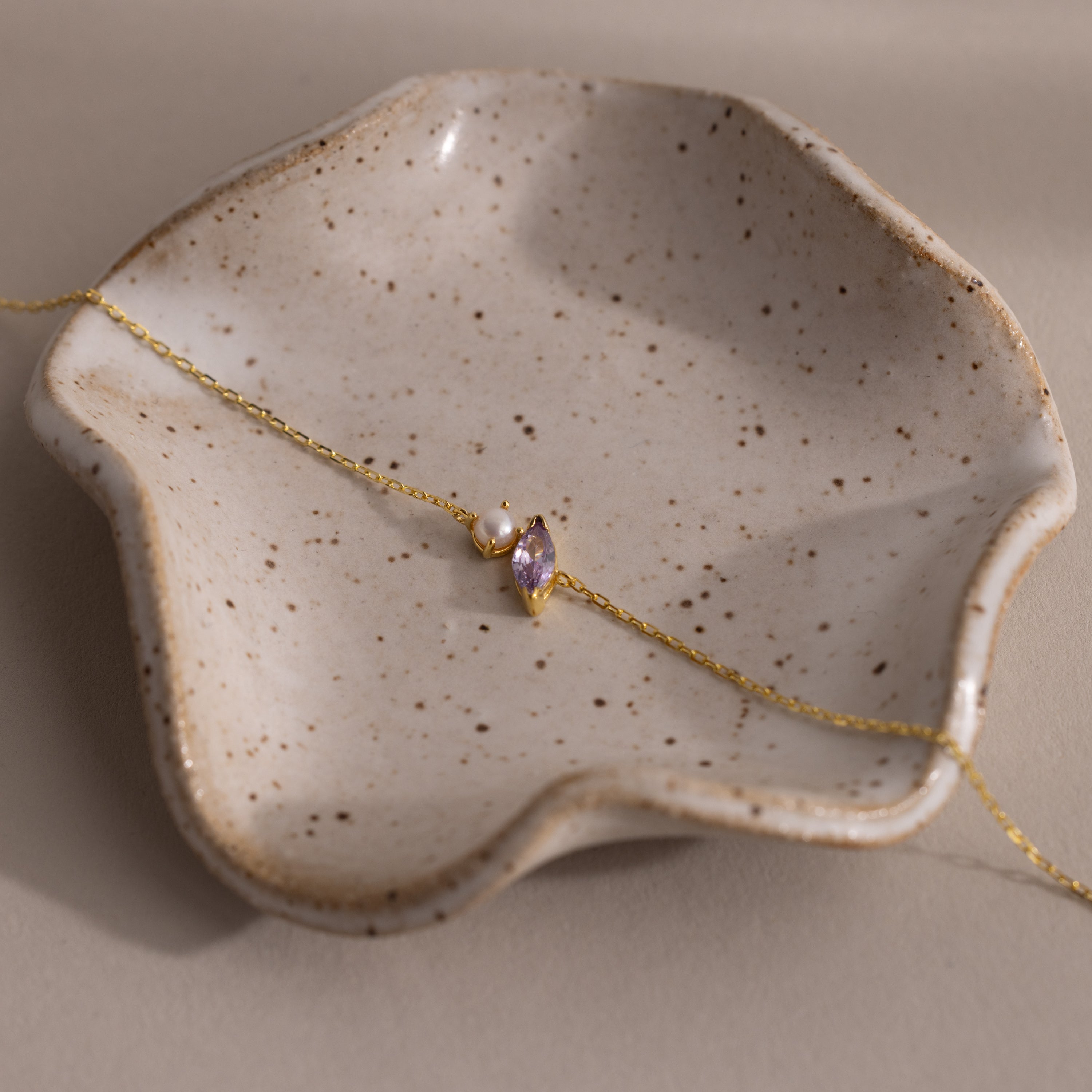 The Marquise Alexandrite & Pearl Bracelet rests on a speckled, wavy ceramic dish—an elegant gift for any special occasion.