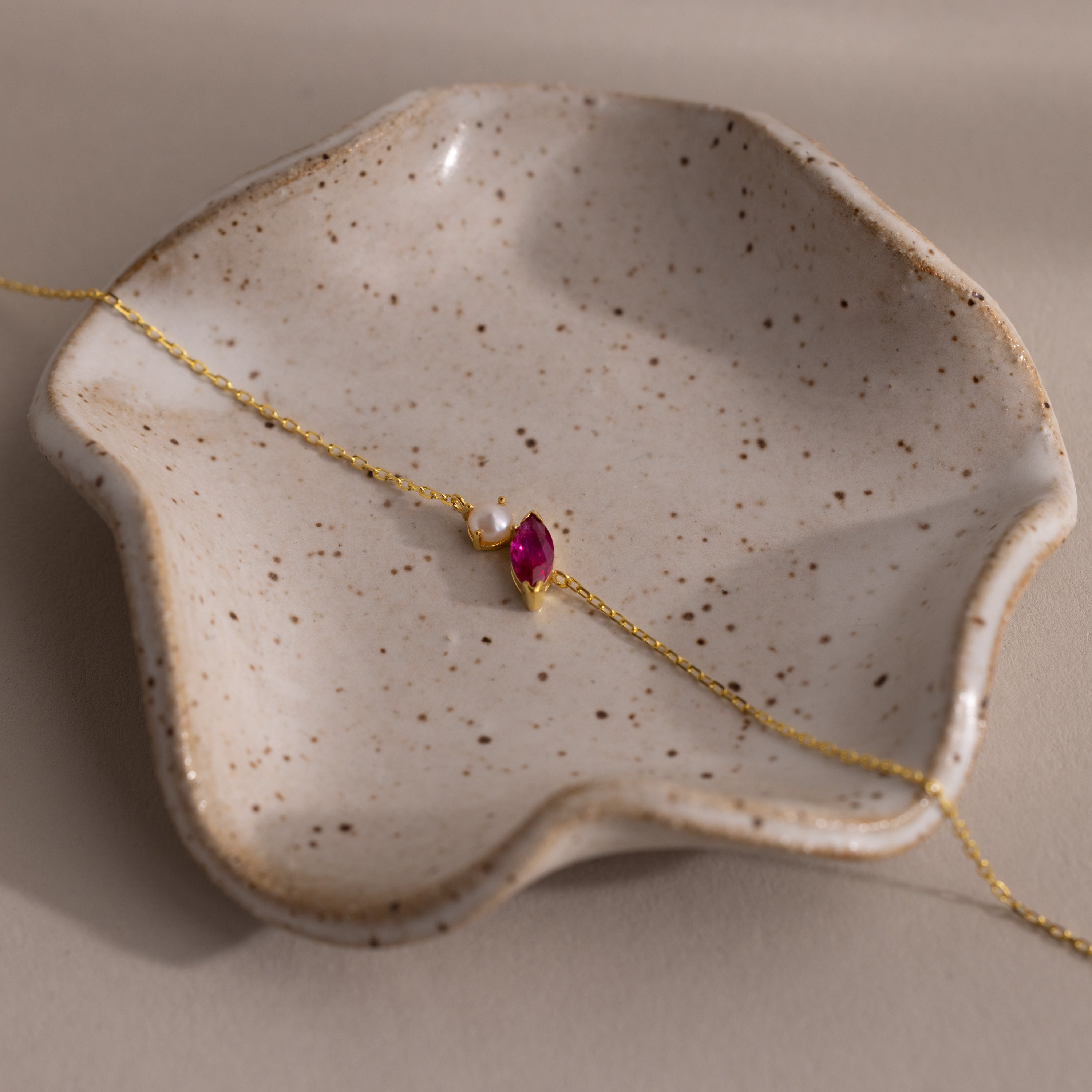 The Marquise Ruby & Pearl Bracelet, featuring a gold chain and pink stone, rests on a speckled ceramic dish—an elegant gift for someone special.