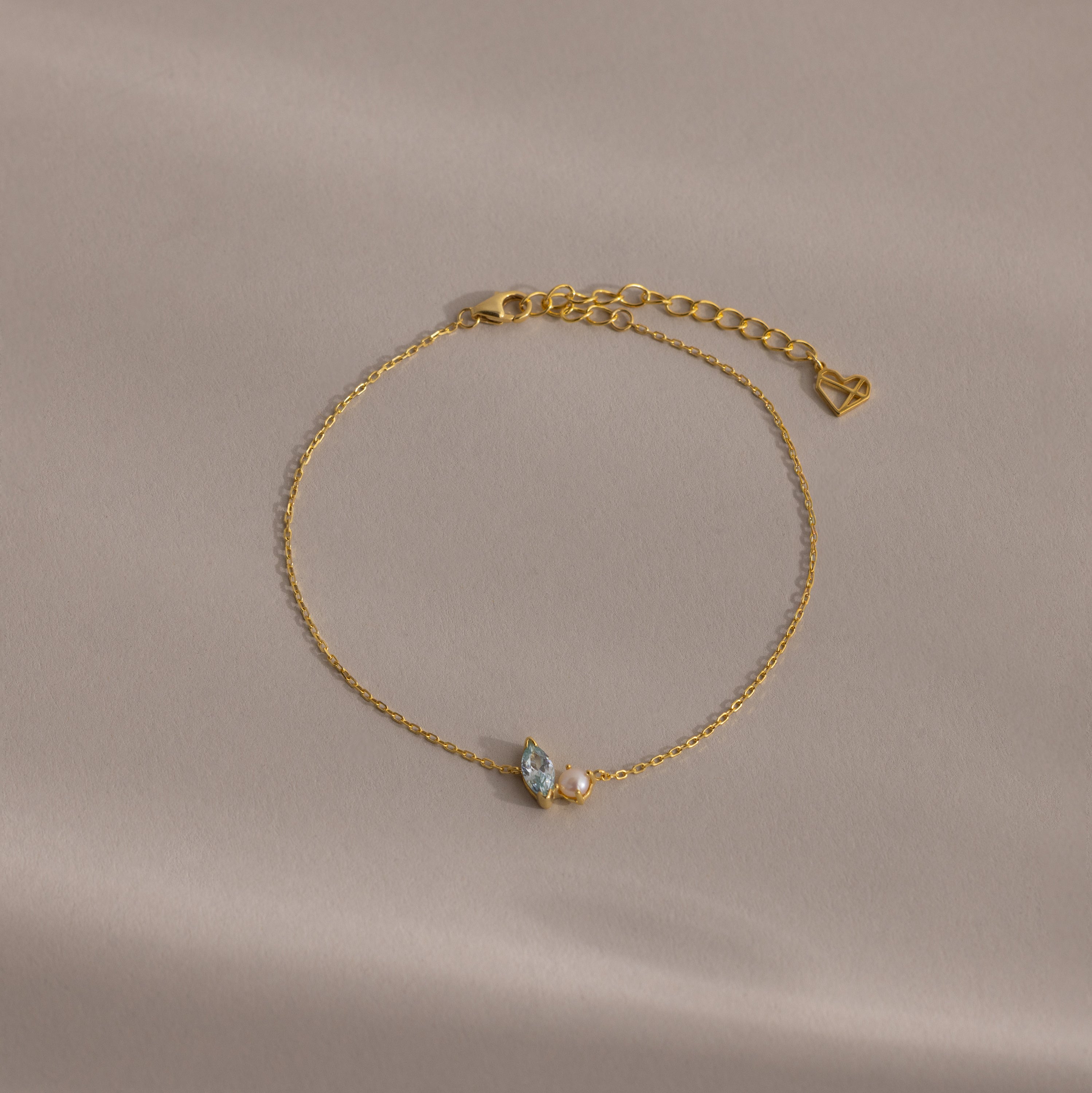 The Marquise Birthstone & Pearl Bracelet features two small gemstones and a heart charm, beautifully displayed on a beige surface—an ideal gift for someone special.