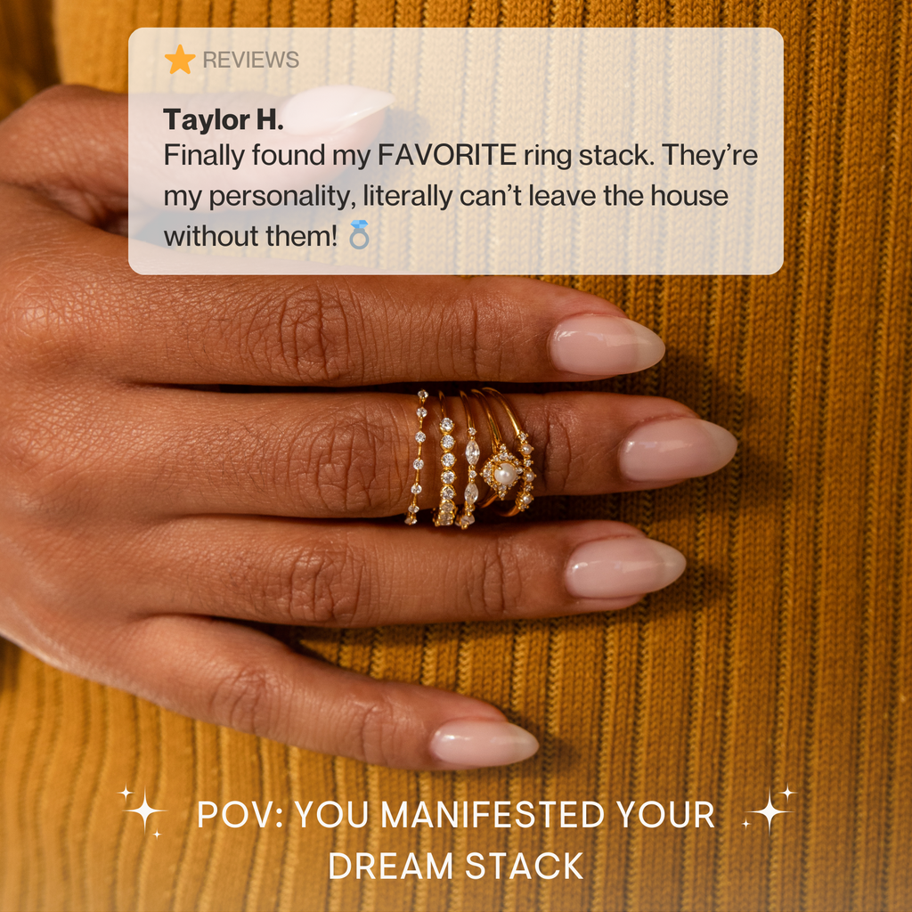 A hand with nude manicured nails wears multiple gold and pearl rings, including the Pearl Diamond Stacking Rings, against a mustard yellow sweater.