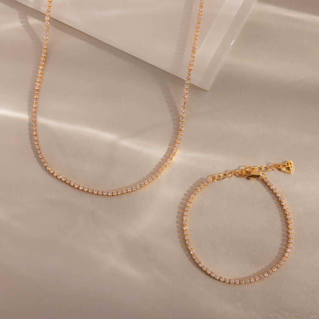 The Dainty Diamond Tennis Necklace & Bracelet Set, adorned with small clear crystals, rests elegantly on beige satin—perfect for lovers of delicate diamond jewelry.
