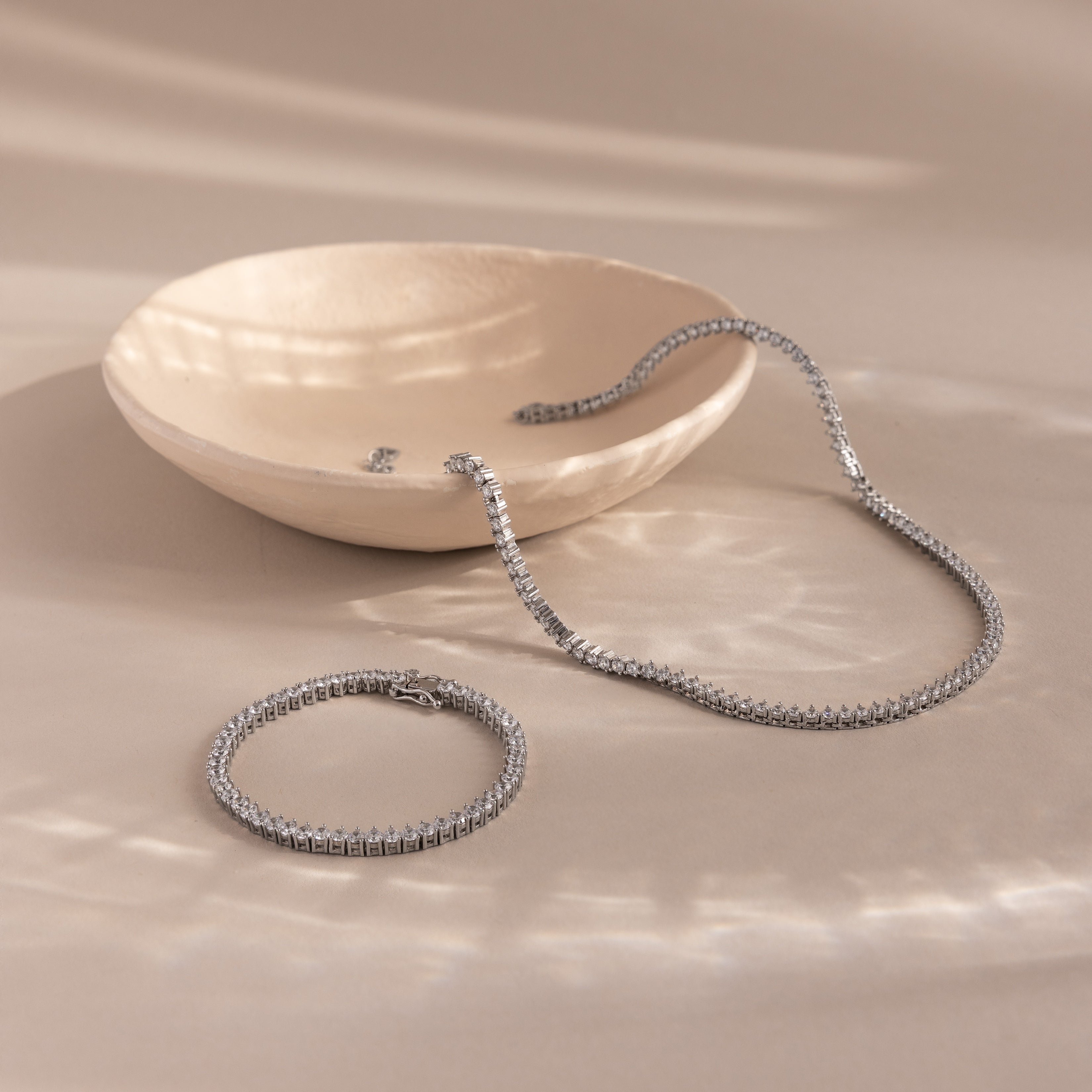 3-Prong Diamond Tennis Necklace & Bracelet Set in White Gold, both with classic 3-prong settings, displayed on a beige surface beside a shallow ceramic dish.
