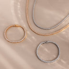 The 3-Prong Diamond Tennis Necklace & Bracelet Set, featuring gold and silver tone pieces with a 3-prong diamond bracelet, is displayed on a beige surface.