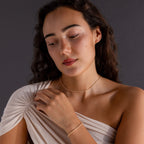 Woman with wavy dark hair wears a cream off-shoulder top and the Dainty Diamond Tennis Necklace & Bracelet Set in 18K Gold—an elegant choice for anniversary jewelry.