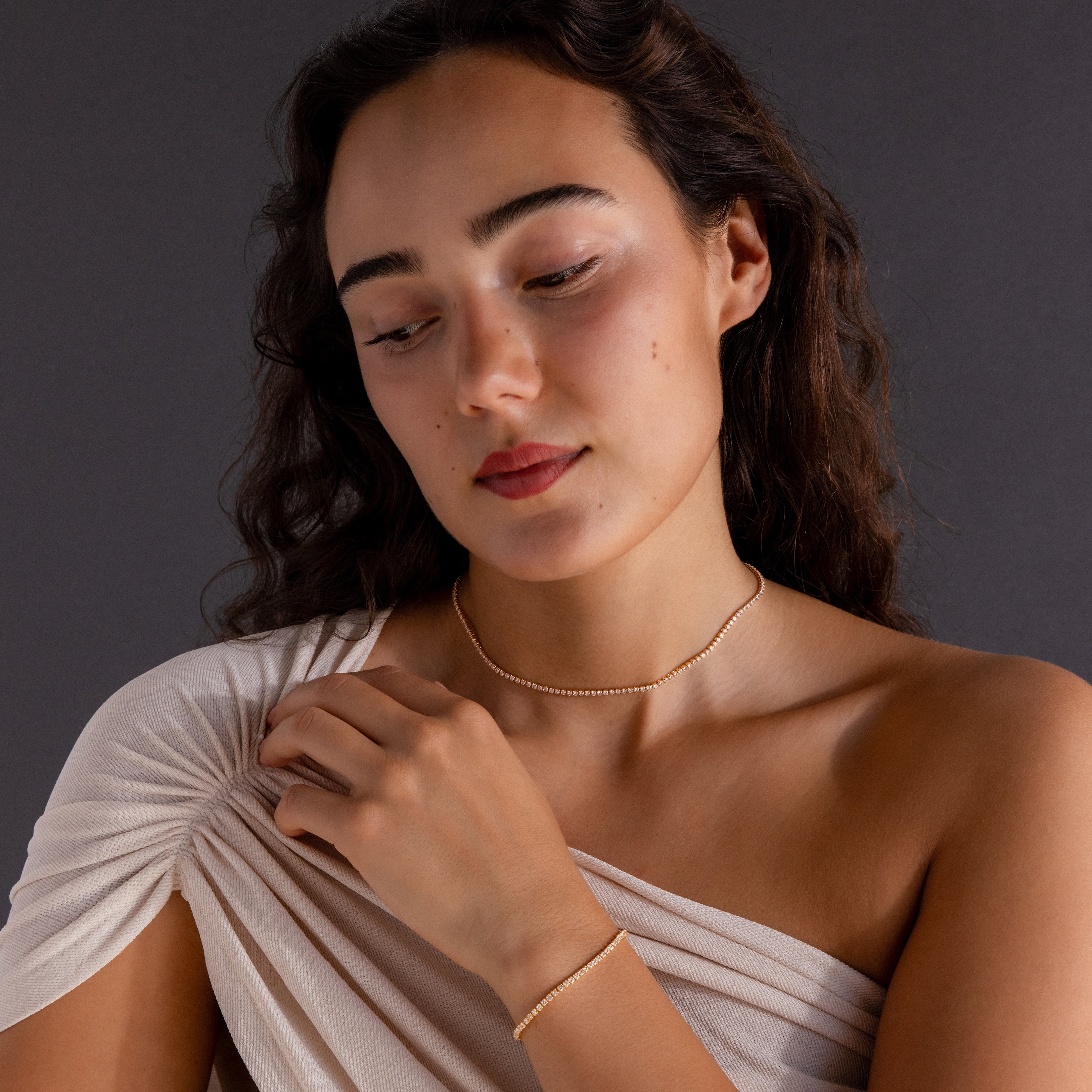 Woman with wavy dark hair wears a cream off-shoulder top and the Dainty Diamond Tennis Necklace & Bracelet Set in 18K Gold—an elegant choice for anniversary jewelry.