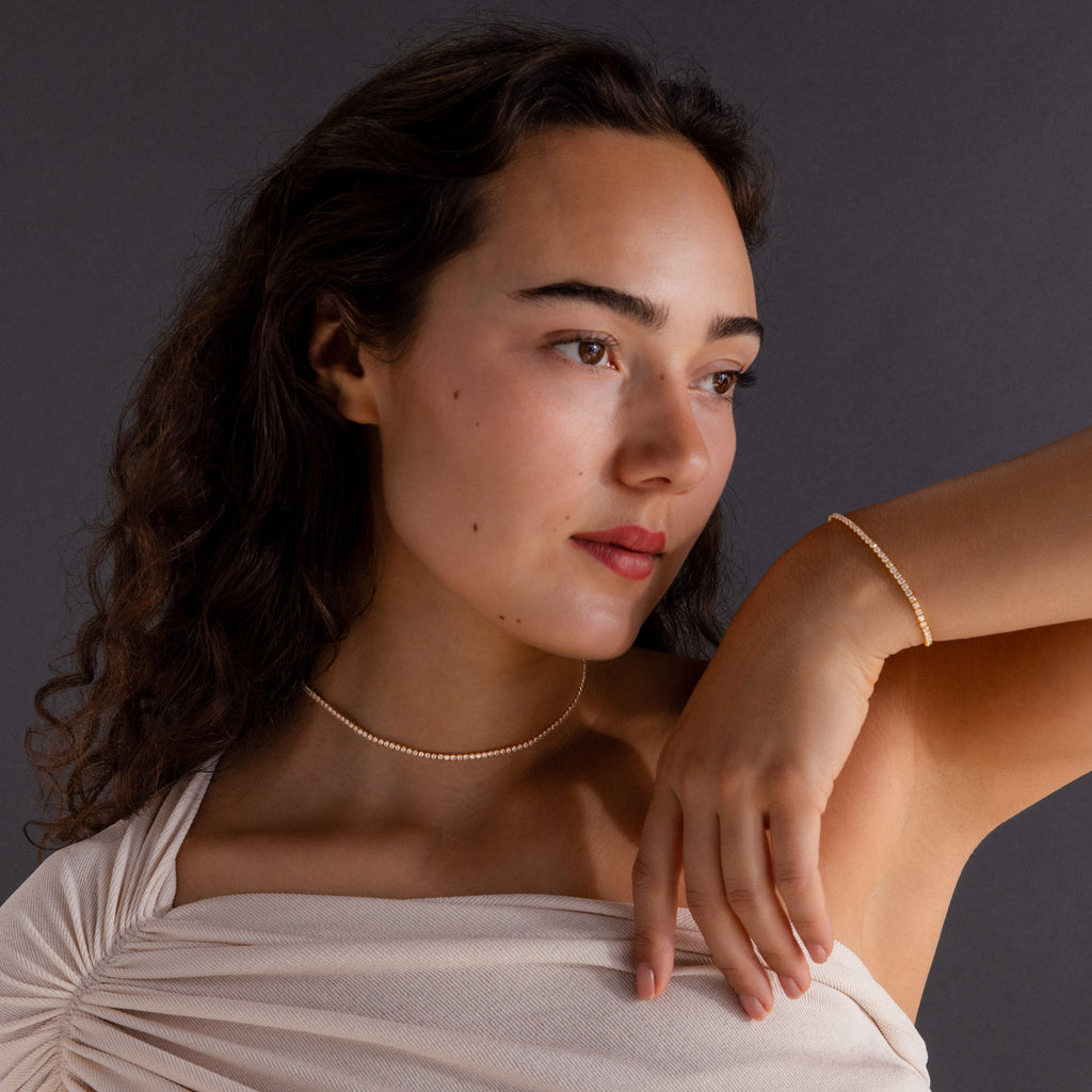 Woman with long curly hair wears a cream off-shoulder top and the Dainty Diamond Tennis Necklace & Bracelet Set, posing thoughtfully.