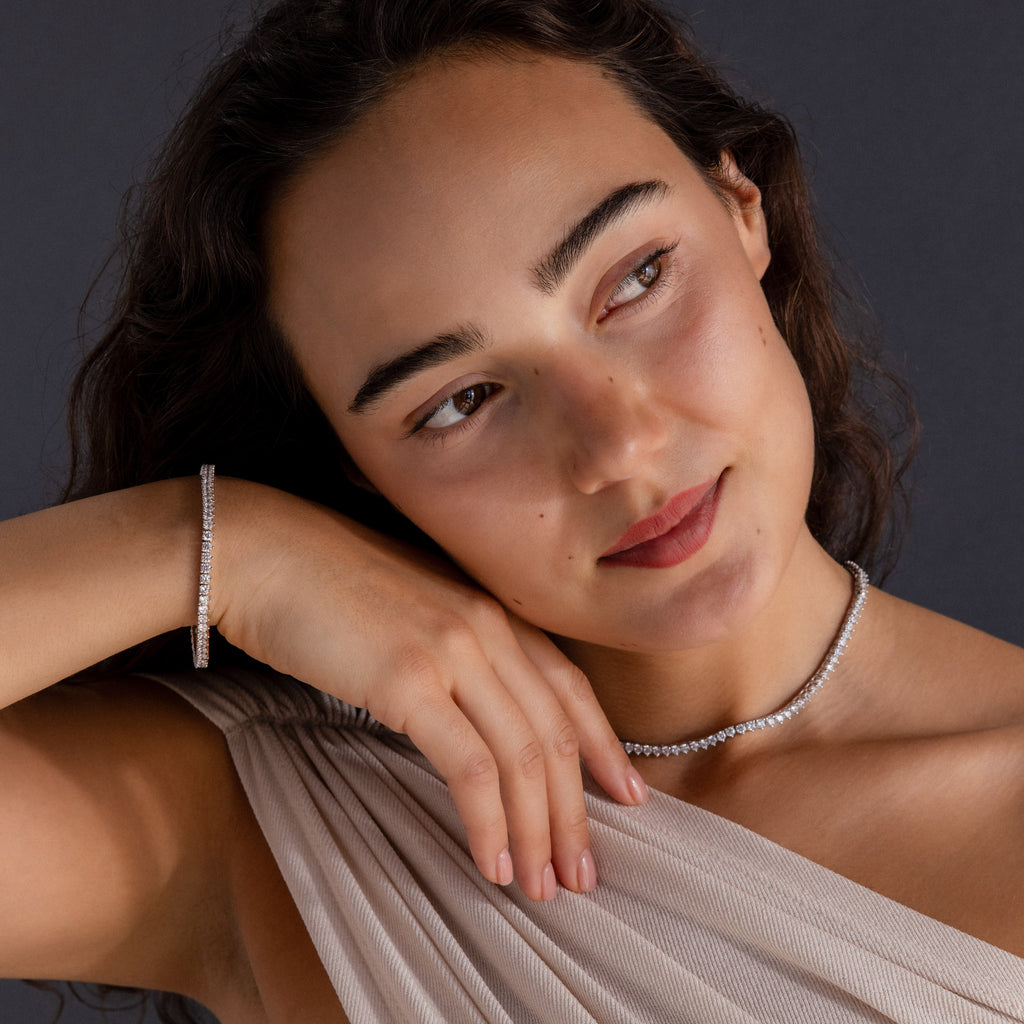 A woman with dark hair, wearing a beige top and gazing to the side, showcases the 3-Prong Diamond Tennis Necklace & Bracelet Set in White Gold.