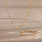 The Dainty Diamond Tennis Necklace & Bracelet Set in 18K Gold sparkles with small diamonds on a neutral beige background—an elegant choice for anniversary jewelry.