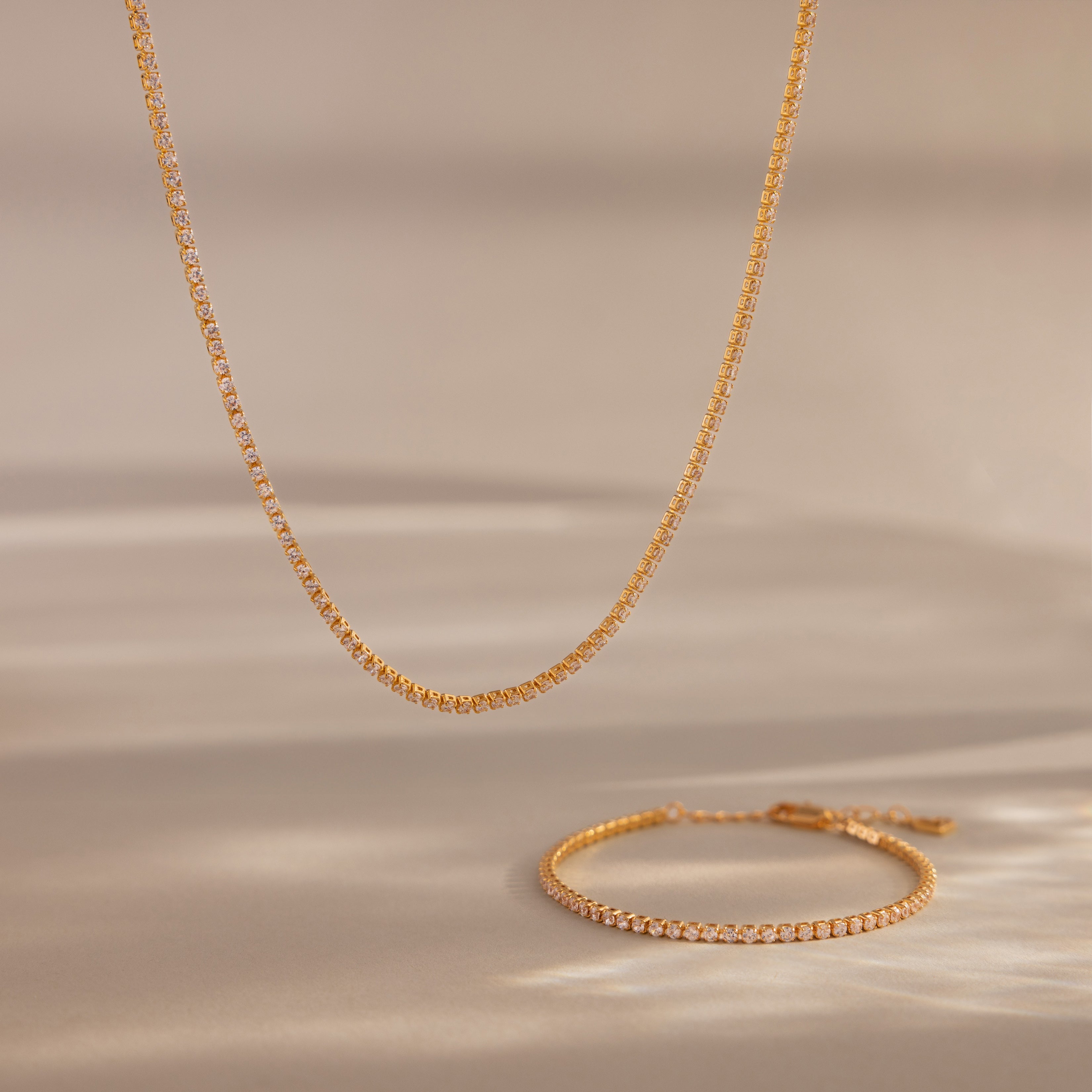 The Dainty Diamond Tennis Necklace & Bracelet Set in 18K Gold sparkles with small diamonds on a neutral beige background—an elegant choice for anniversary jewelry.