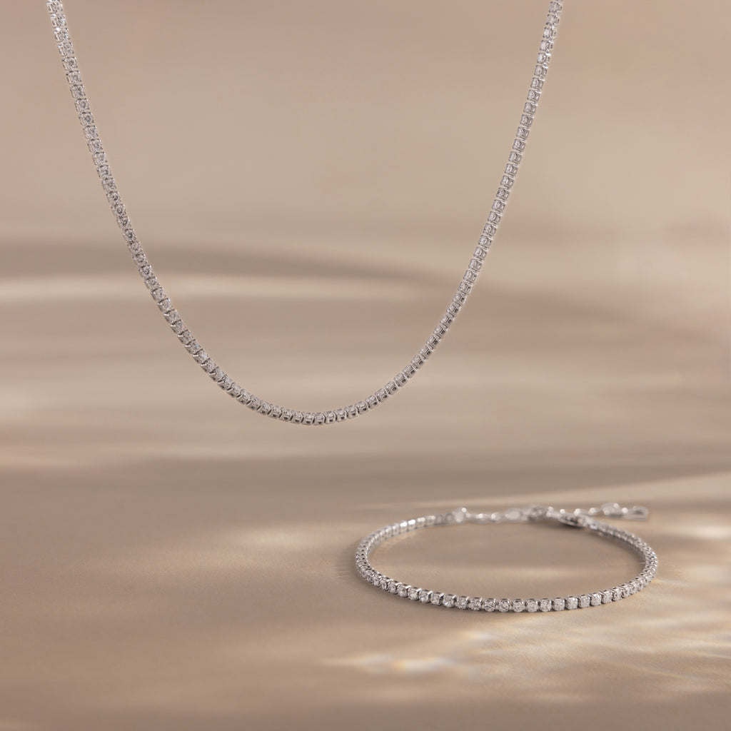The Dainty Diamond Tennis Necklace & Bracelet Set in White Gold, featuring bezel-set diamonds, is displayed on a beige surface with soft lighting.