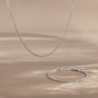 The Dainty Diamond Tennis Necklace & Bracelet Set in White Gold, featuring bezel-set diamonds, is displayed on a beige surface with soft lighting.