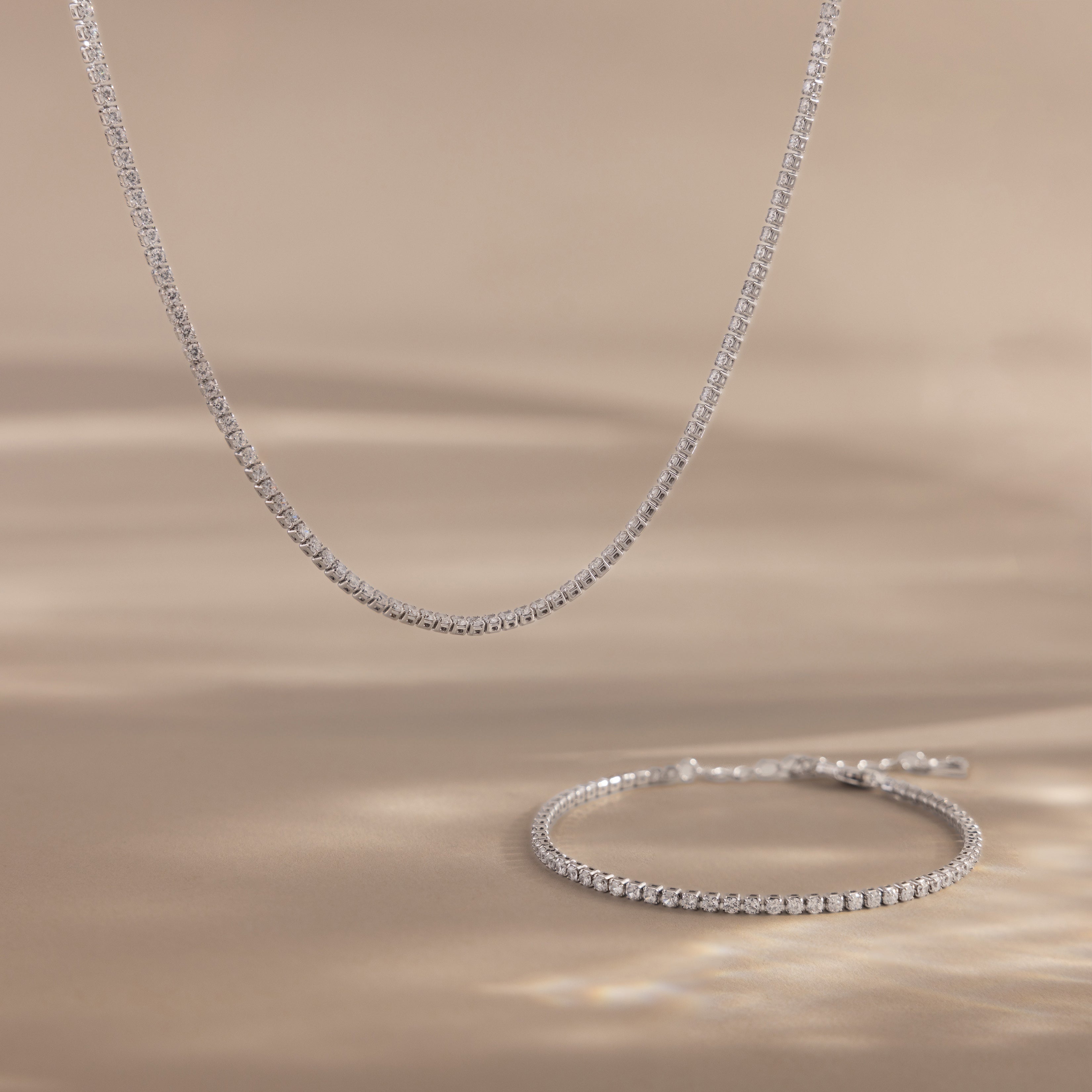 The Dainty Diamond Tennis Necklace & Bracelet Set in White Gold, featuring bezel-set diamonds, is displayed on a beige surface with soft lighting.