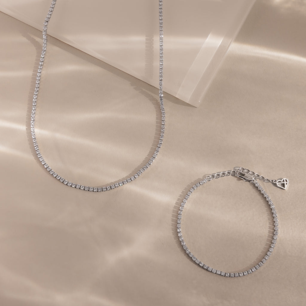 The Dainty Diamond Tennis Necklace & Bracelet Set in White Gold is shown on a beige, sunlit surface with soft shadows, highlighting the bezel-set diamonds.