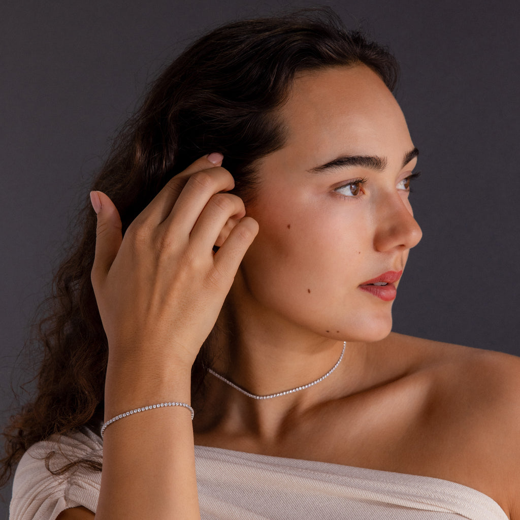 Woman with long hair wears the Dainty Diamond Tennis Necklace & Bracelet Set, gazing to the side against a dark background.
