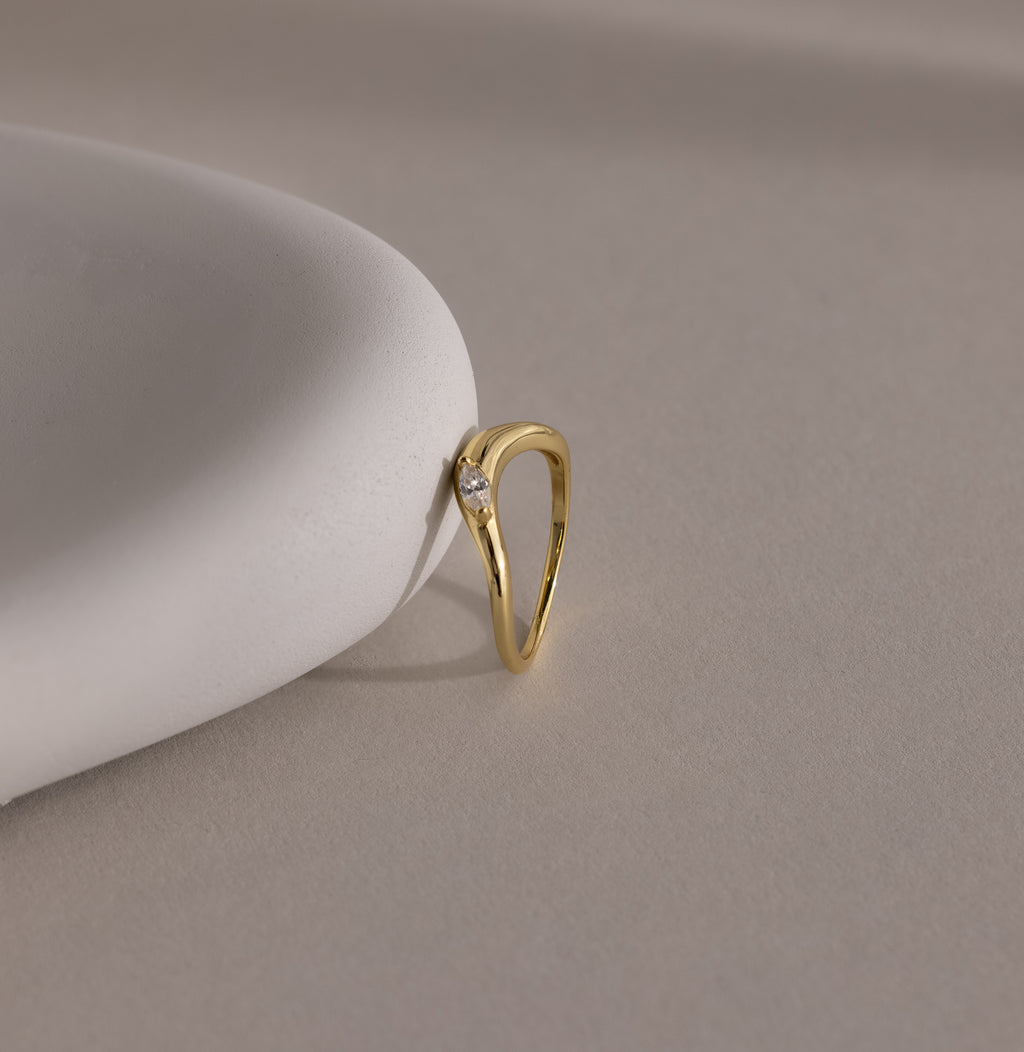 The Zen Diamond Curve Ring, crafted in gold and featuring a single marquise diamond accent, is displayed on a smooth, white surface with a neutral background.