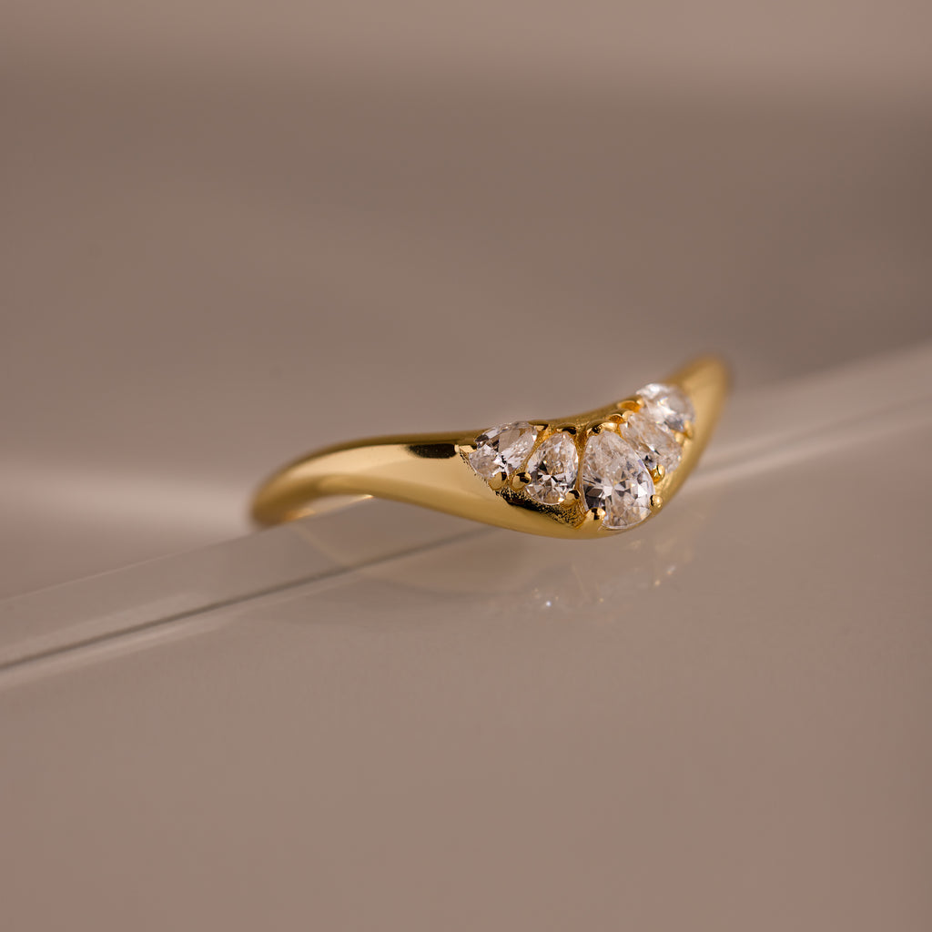 Gold curve ring with tapered band featuring five pear cut diamonds in a graduated V shaped setting, displayed on a neutral glass background.