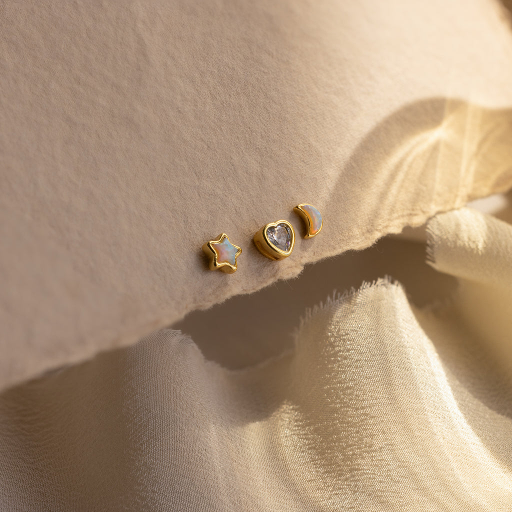 Opal star, CZ diamond heart, and opal moon stud earrings in gold displayed up close on a beige background.