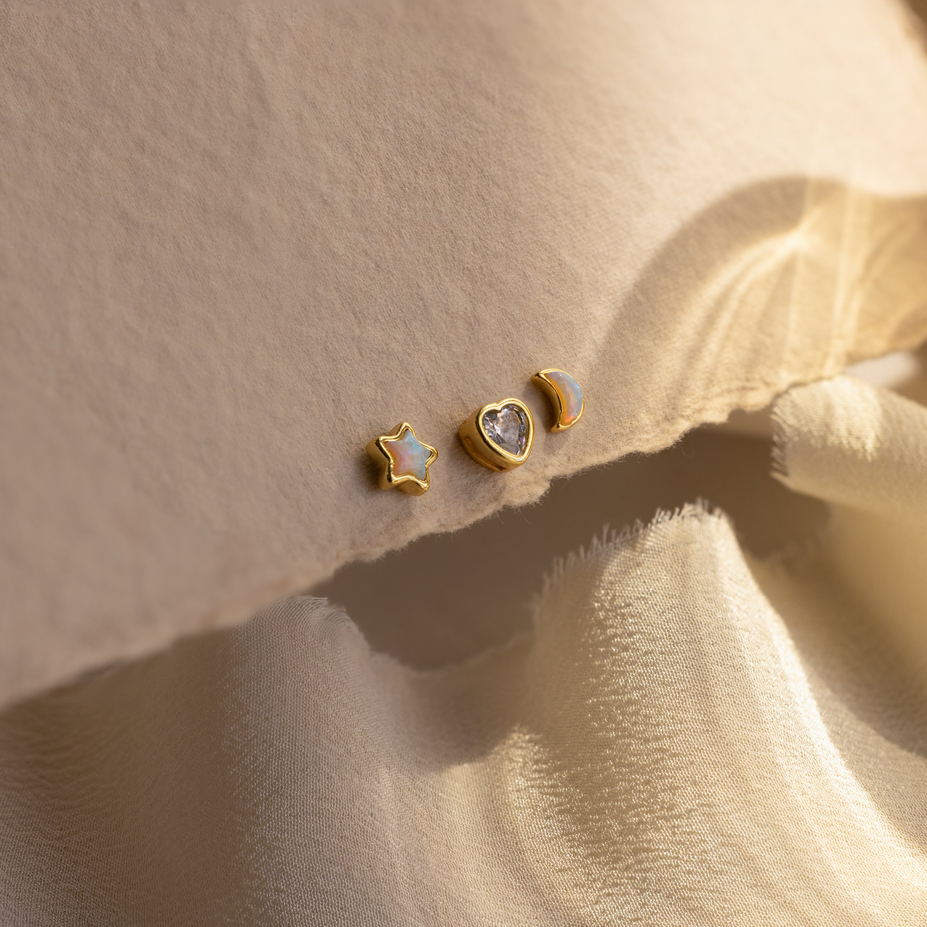 Opal star, CZ diamond heart, and opal moon stud earrings in gold displayed up close on a beige background.