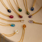 Gold chains with colorful gemstone pendants, including the North Star Birthstone Signet Necklace, are artfully arranged in a circle on a cream-colored surface.