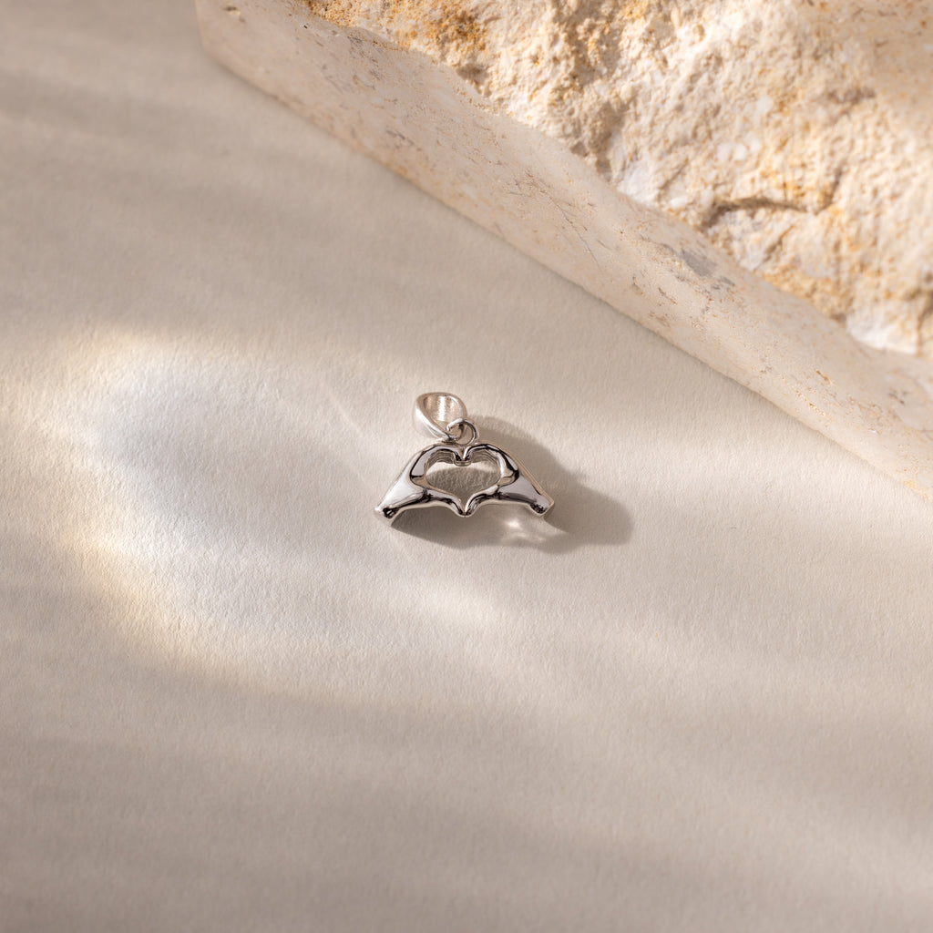 A close up of a silver Heart Hands charm placed on a light surface beside a stone.