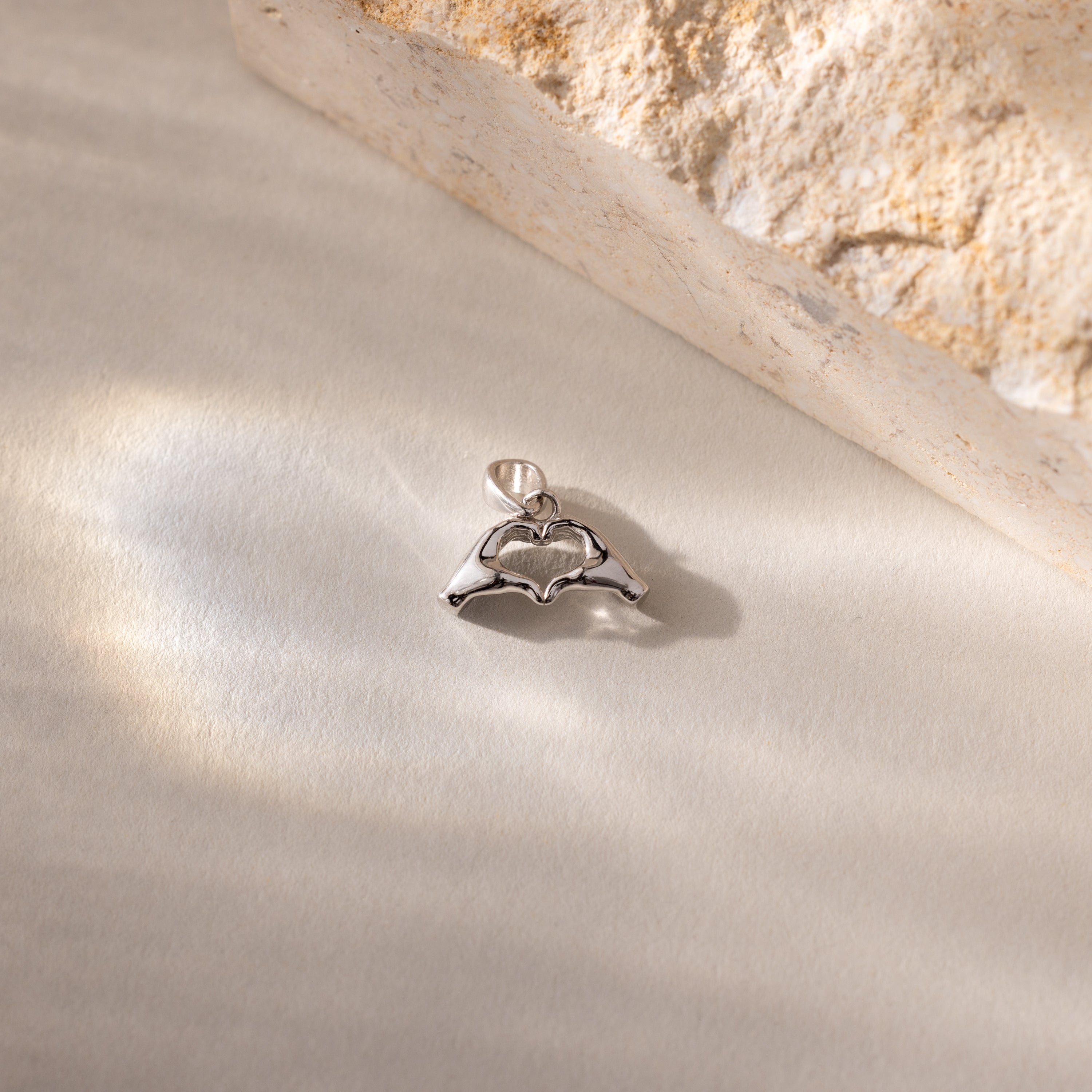 A close up of a silver Heart Hands charm placed on a light surface beside a stone.