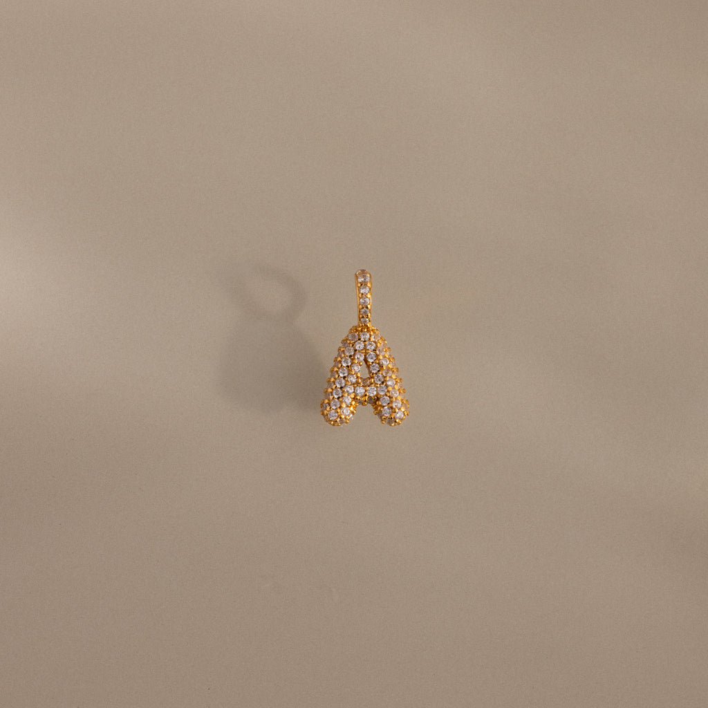 The Pave Bubble Letter Charm features diamond-studded lettering in the shape of an A, displayed on a plain beige background with a subtle shadow.